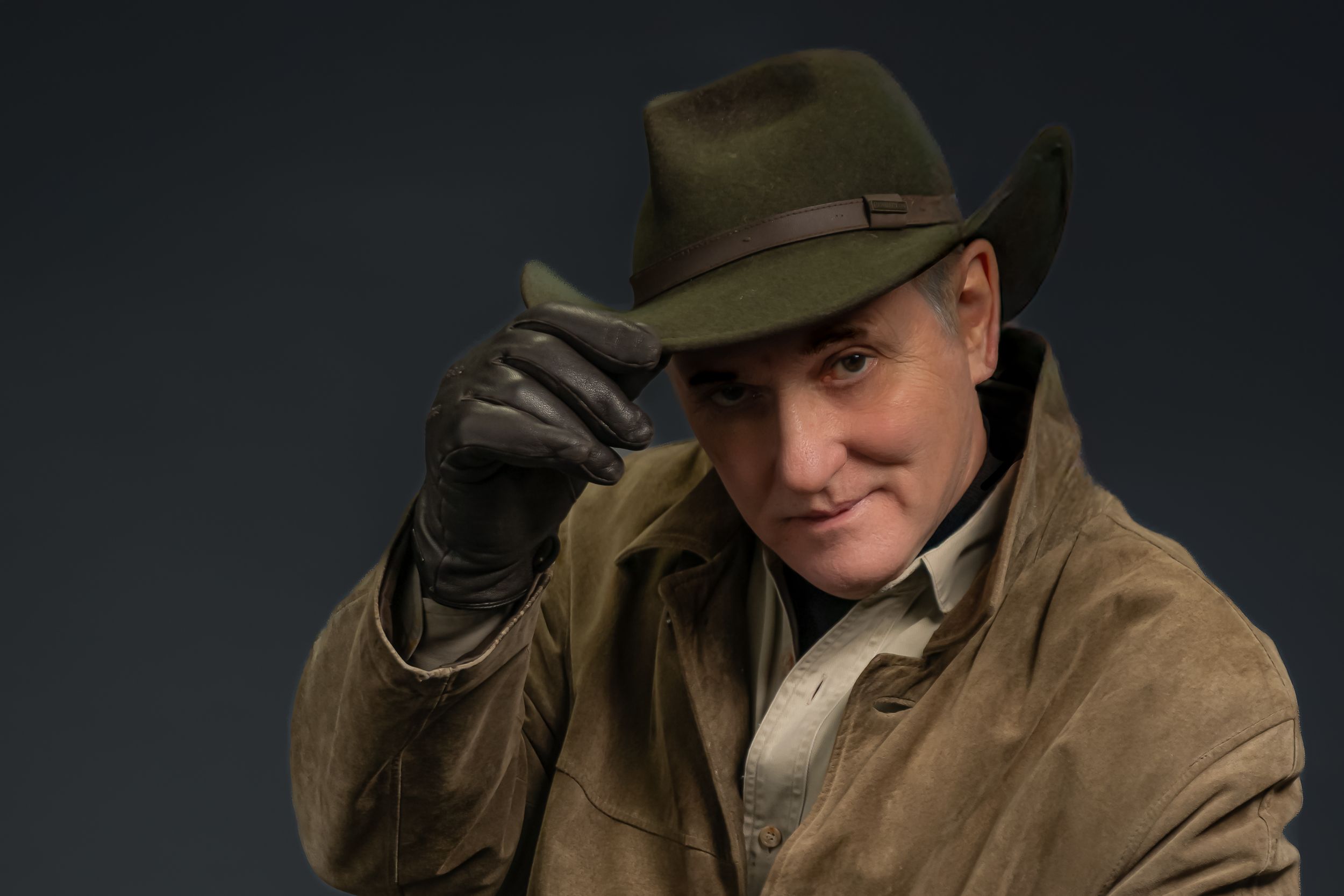 Figure wearing a brown fedora hat and leather jacket tips the brim of their hat in a dramatic lighting setup.