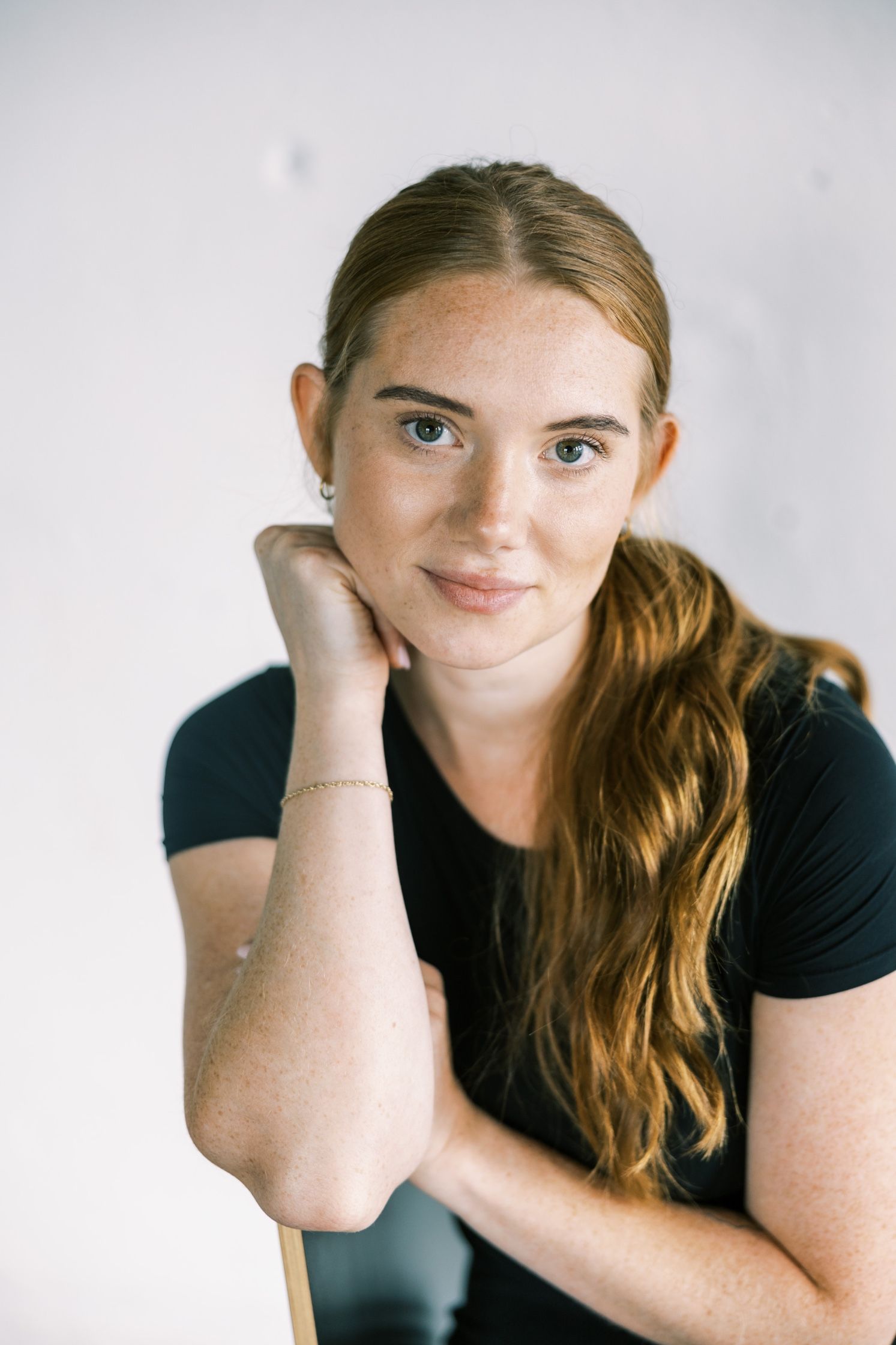 Portrait against white background showing a person with long wavy red hair wearing a black t-shirt in natural lighting.