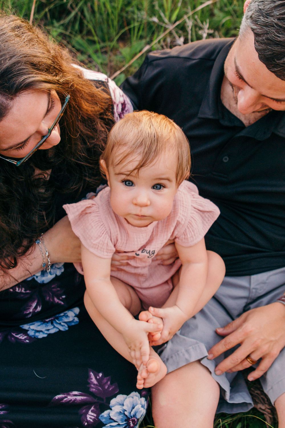 Baby in pink dress sits with adults outdoors on floral blanket during family photo session.