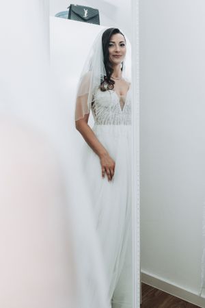 Elegant bride in fitted white gown poses against white wall with veil flowing.