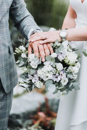 Close-up of newlyweds' hands with rings resting on white floral bouquet.