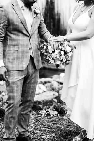 Black and white wedding photo showing clasped hands with bride holding floral bouquet and groom in plaid suit.