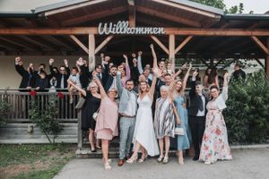 Wedding party celebrates joyfully outside rustic wooden venue with 'Willkommen' sign.