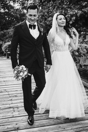 Couple in wedding attire walking together on a wooden boardwalk with the bride holding a floral bouquet.