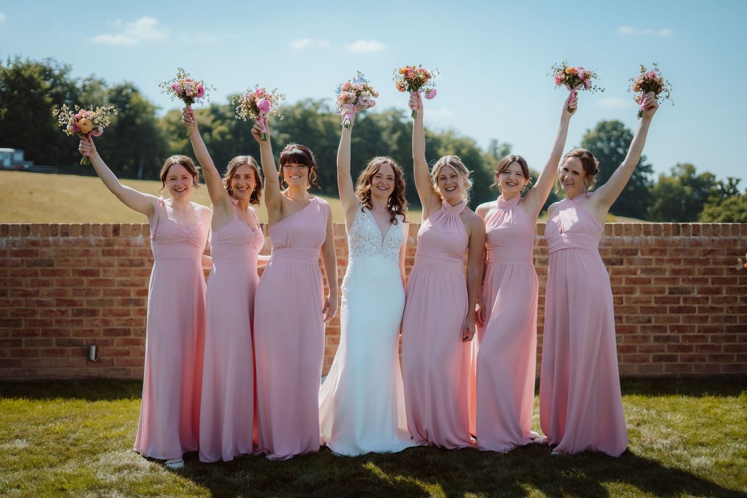 Joyful wedding party in pink dresses and light suits celebrates outdoors on a garden path.