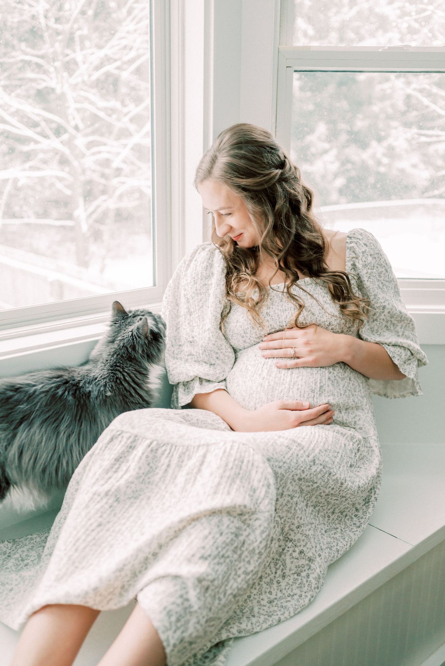 A pregnant woman in a white lace dress sits by a window with a fluffy grey cat while cradling her baby bump.