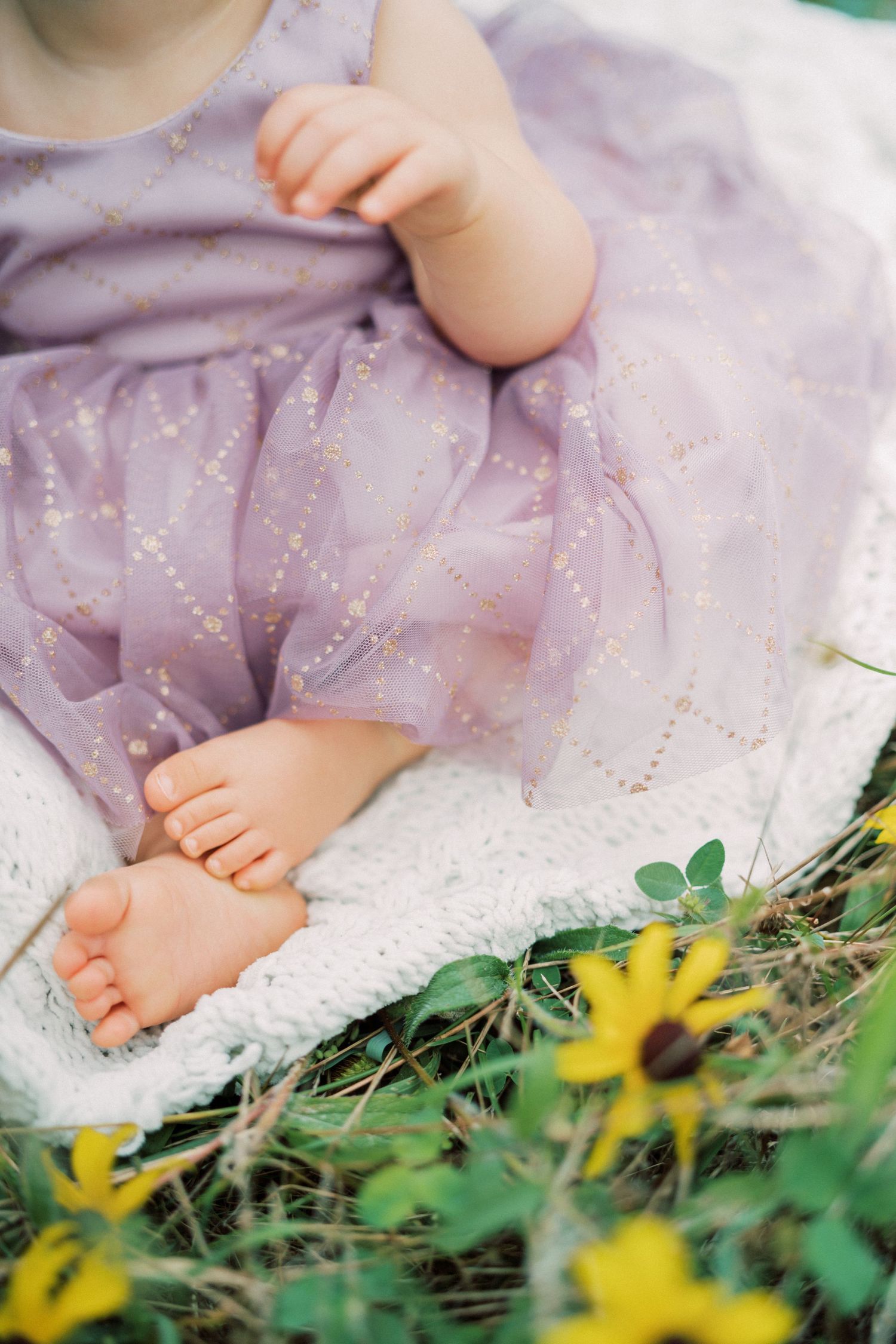 Close-up of baby feet and purple sparkly dress hem with yellow flowers in background.