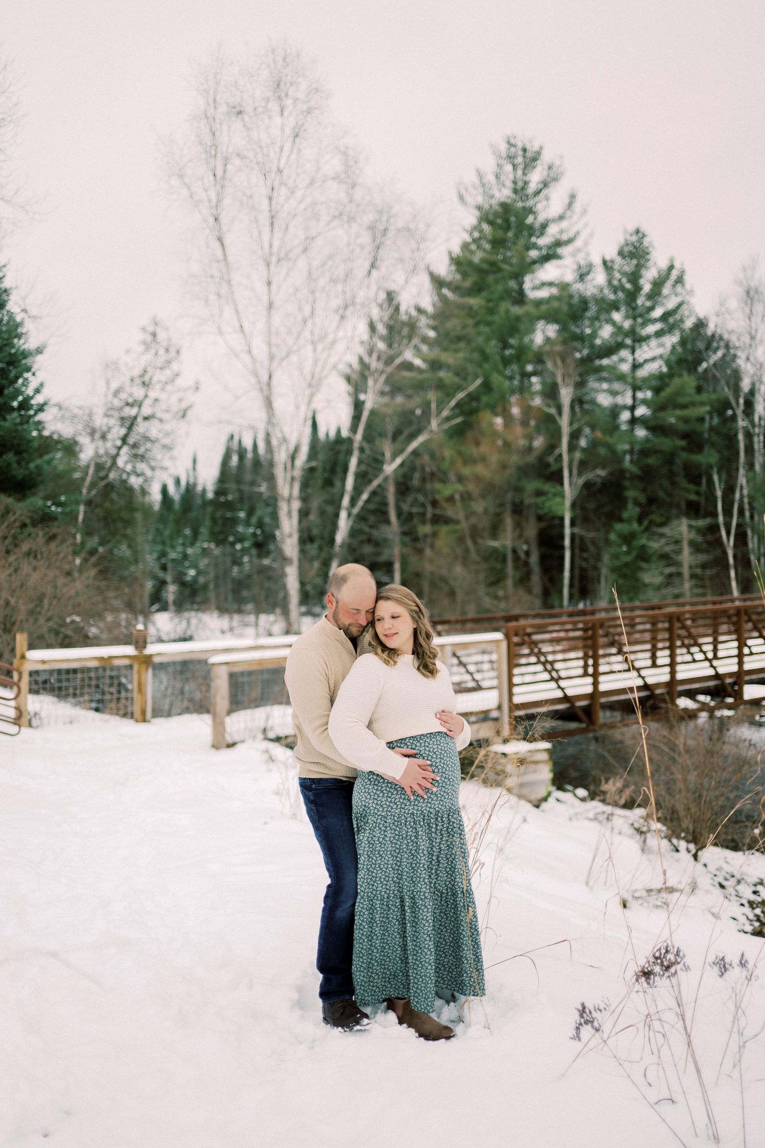 Winter maternity portrait in front of snowy wooden bridge with pine trees in background.
