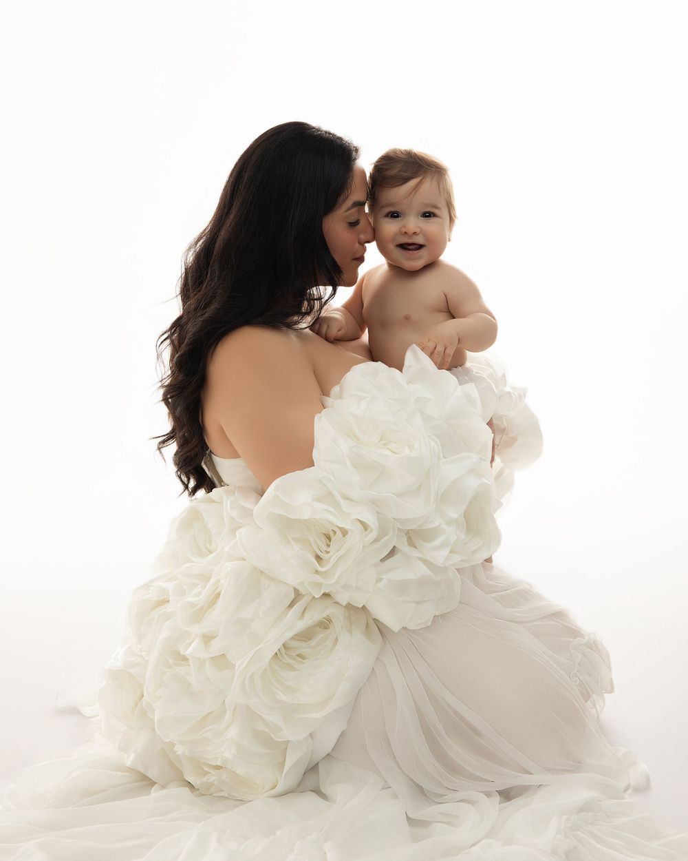 A parent in a flowing white gown holds a joyful baby against a bright white studio backdrop.