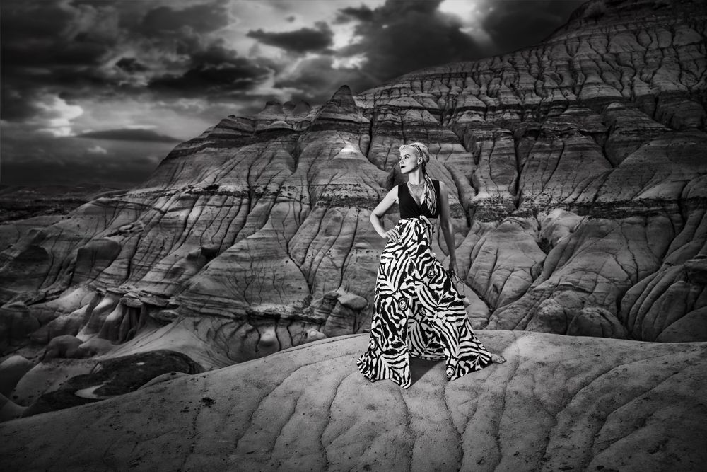 A figure in flowing patterned dress stands dramatically atop eroded rock formations under dramatic stormy sky. Drumheller, Alberta.