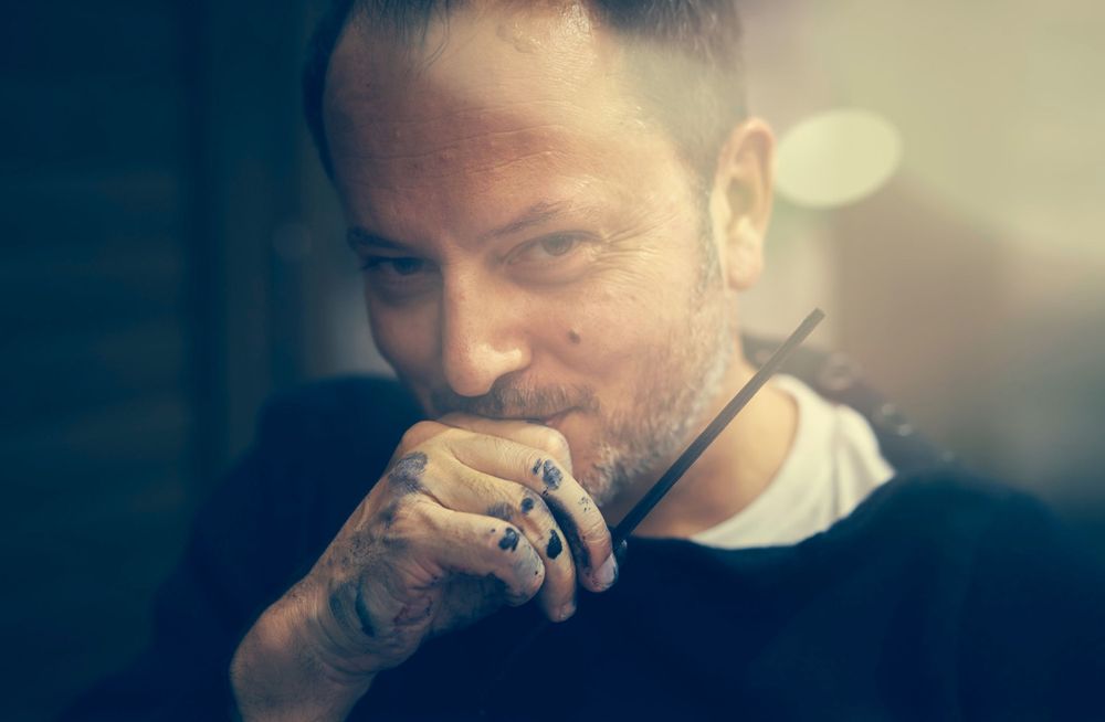 Nicolas Lefeuvre in dark sweater after finishing a painting with paint on his hands and soft lighting and bokeh effect in background.