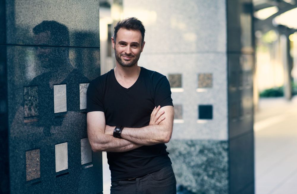 Confident person in black v-neck t-shirt stands with arms crossed against an urban wall with a warm smile - Yves Dalbiez