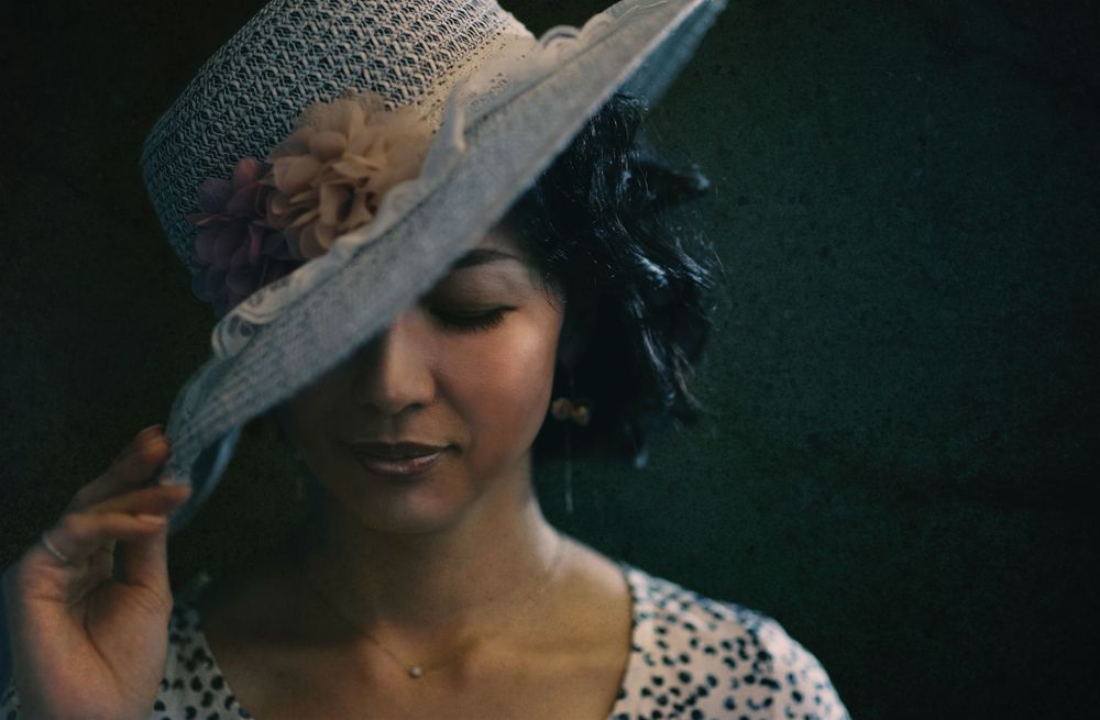 A figure in a decorative sun hat with flower detail smiles gently against a dark background.