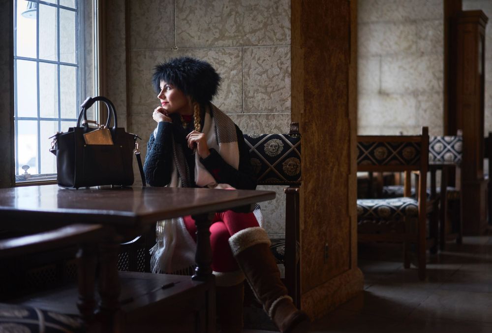 A woman sitting alone at a wooden table in a moody restaurant interior with leather seating and large windows.