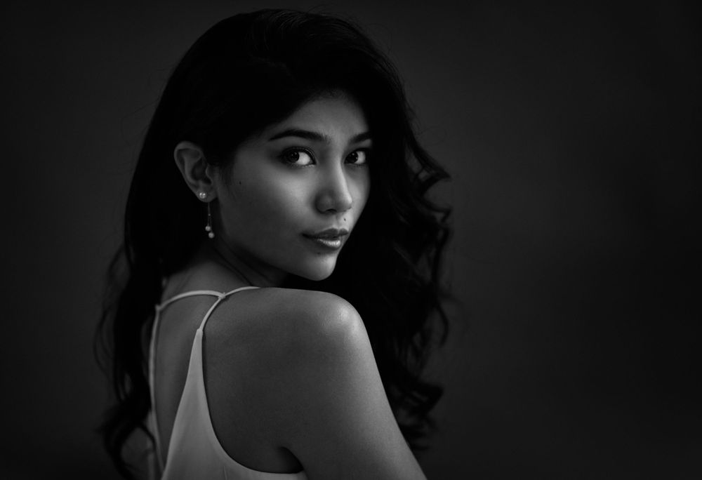 Black and white portrait of Priyanka Yoshikawa looking over shoulder with elegant makeup and dark wavy hair.
