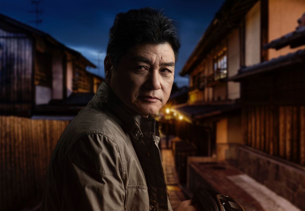 A person in a brown jacket stands in a traditional Japanese alleyway at dusk with wooden buildings illuminated behind them.