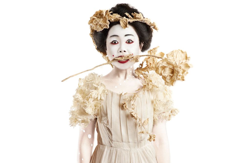 A whimsical figure in vintage white dress with ruffles and theatrical makeup holds a dried flower stem in her mouth in studio on white back