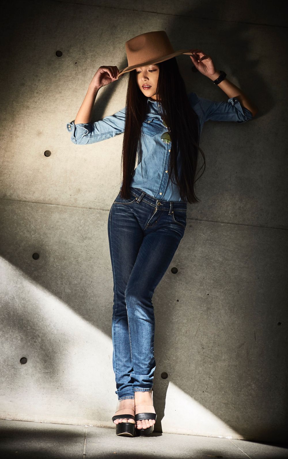 Maki Shima in denim double jeans and brown hat poses stylishly against a concrete wall in dramatic lighting in Tokyo