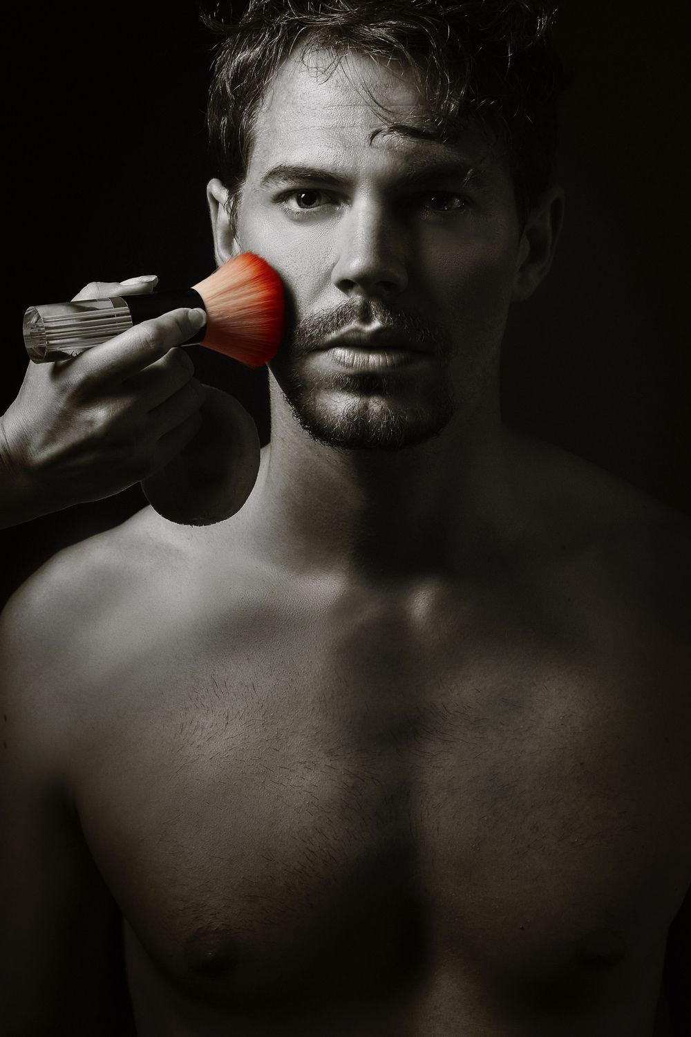 Fine makeup brush softly grazes cheek in dramatic black and white portrait of Dean Newcombe against dark background.