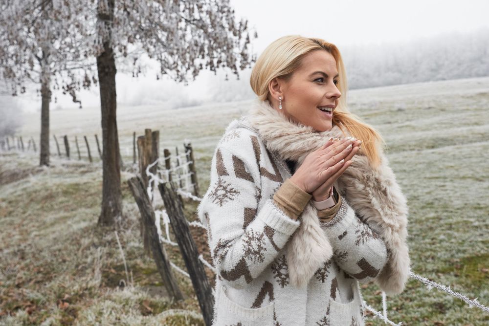 A woman in a cozy winter sweater and fur scarf stands in a frosty landscape with bare trees on a cold morning in France.