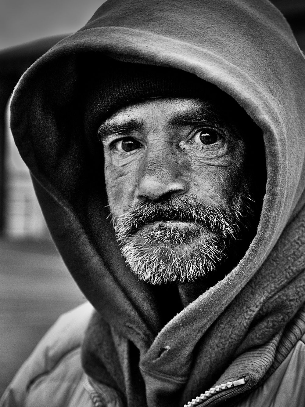A dramatic black and white portrait of someone wearing a hooded sweatshirt with intense gaze and weathered beard in New York Coney Island