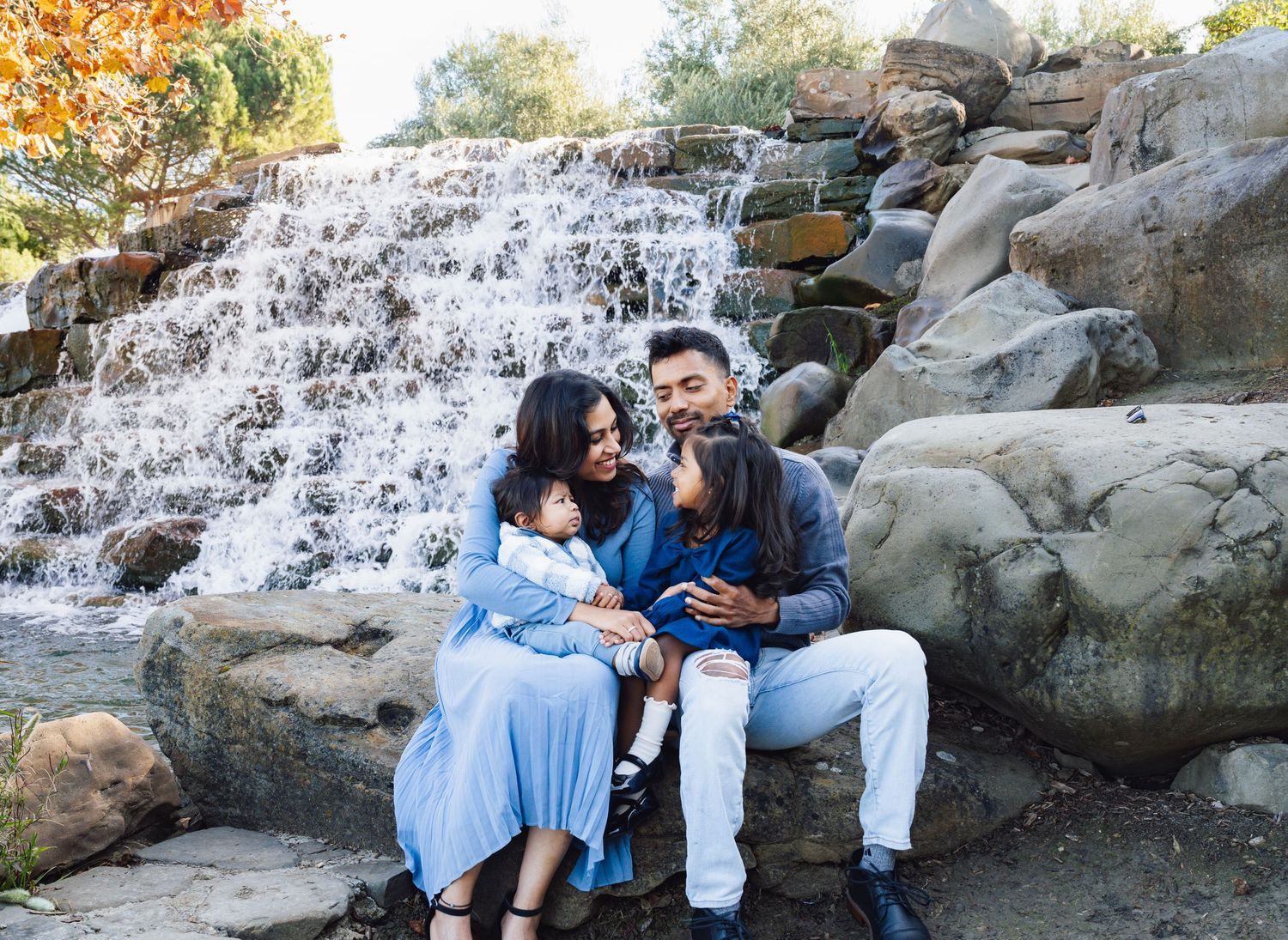 A couple sits together on rocks near a cascading waterfall on an autumn day.