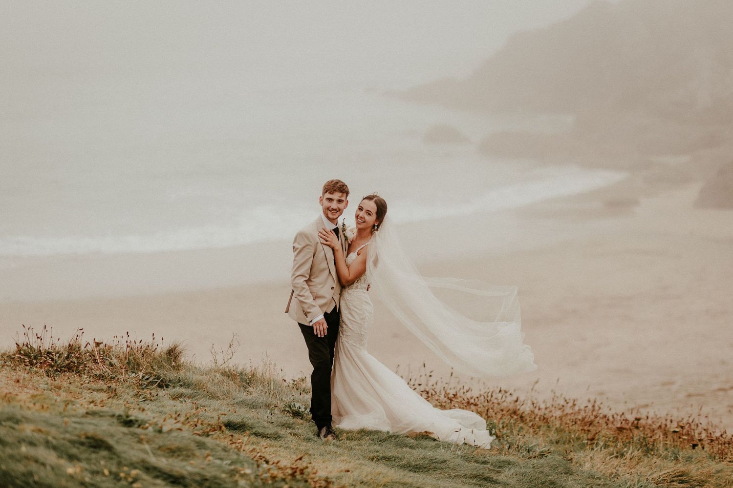 A romantic wedding couple embraces on a misty hilltop at sunset with a flowing veil in the wind.