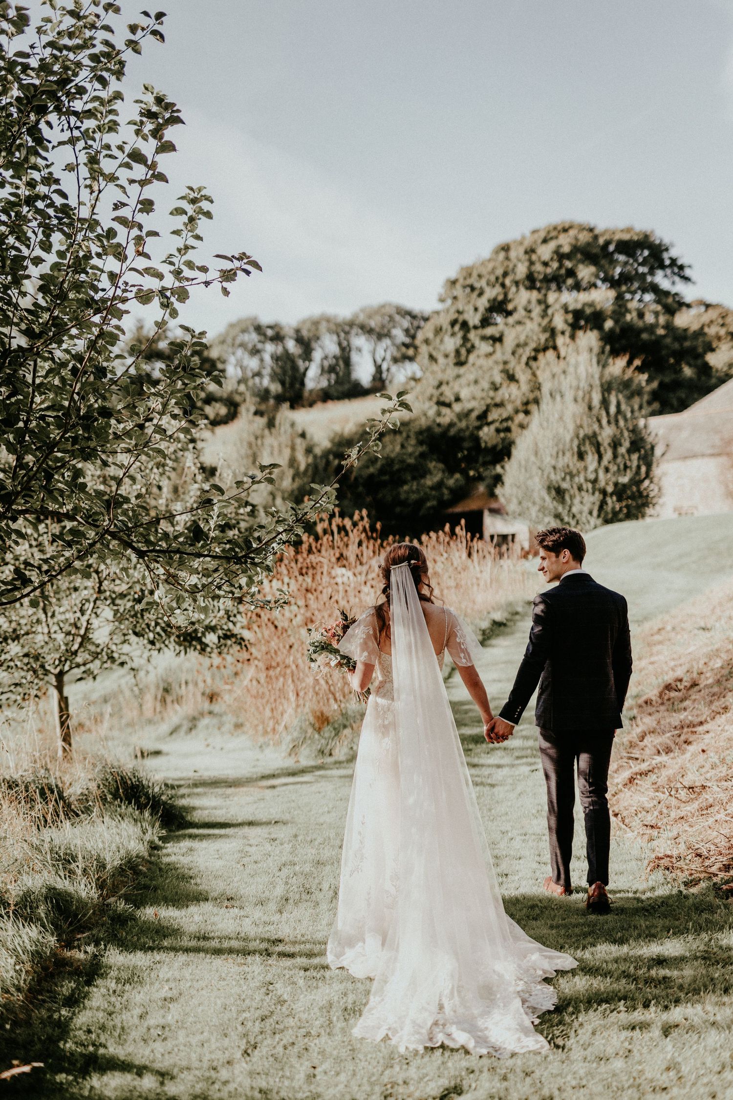 A couple walking hand in hand along a countryside path, with the bride wearing a flowing white veil and dress.