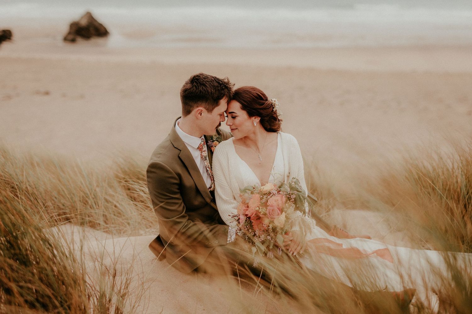 A romantic wedding couple sits together in tall grass on the beach at sunset holding a bouquet of flowers.