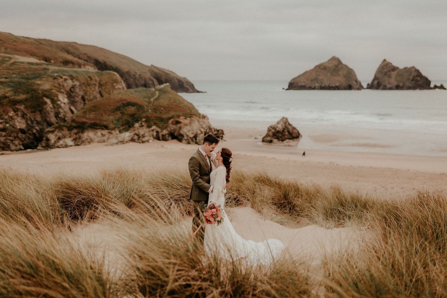 Two figures in wedding attire stand among coastal grass dunes overlooking a dramatic rocky beach and ocean landscape.