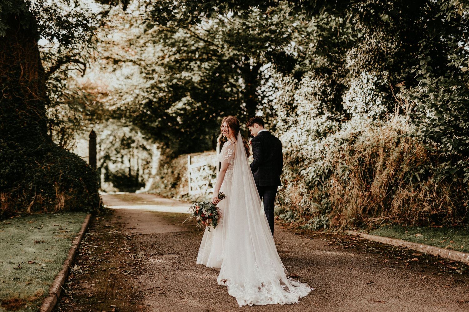 Couple stands together along a tree-lined garden path at golden hour in elegant wedding attire.