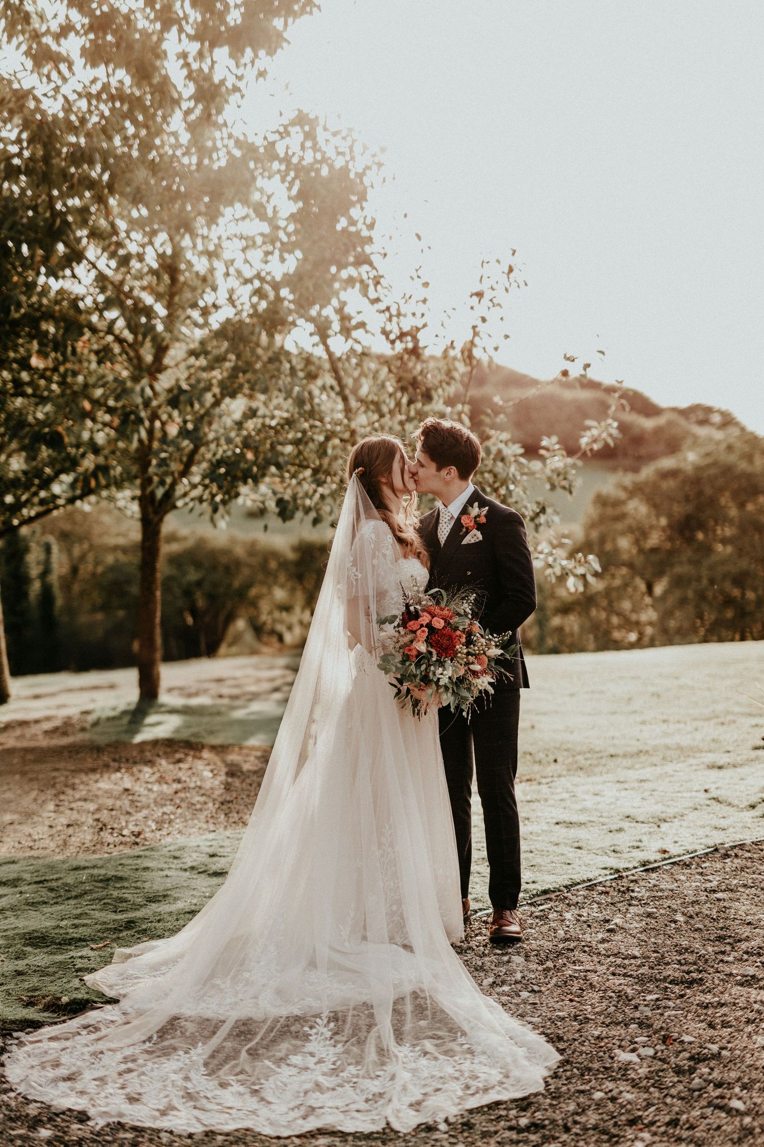 A romantic outdoor wedding photo featuring a couple embracing in warm sunlight with a dramatic trailing lace veil and red bouquet.