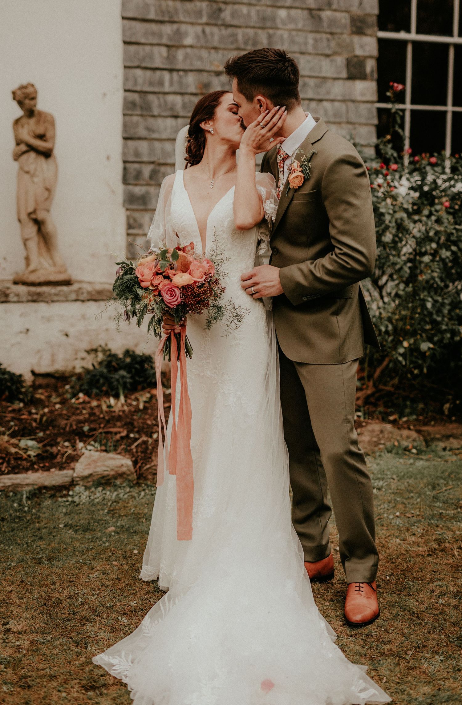 A romantic wedding couple shares an intimate moment in front of a stone building with a classical garden statue nearby.