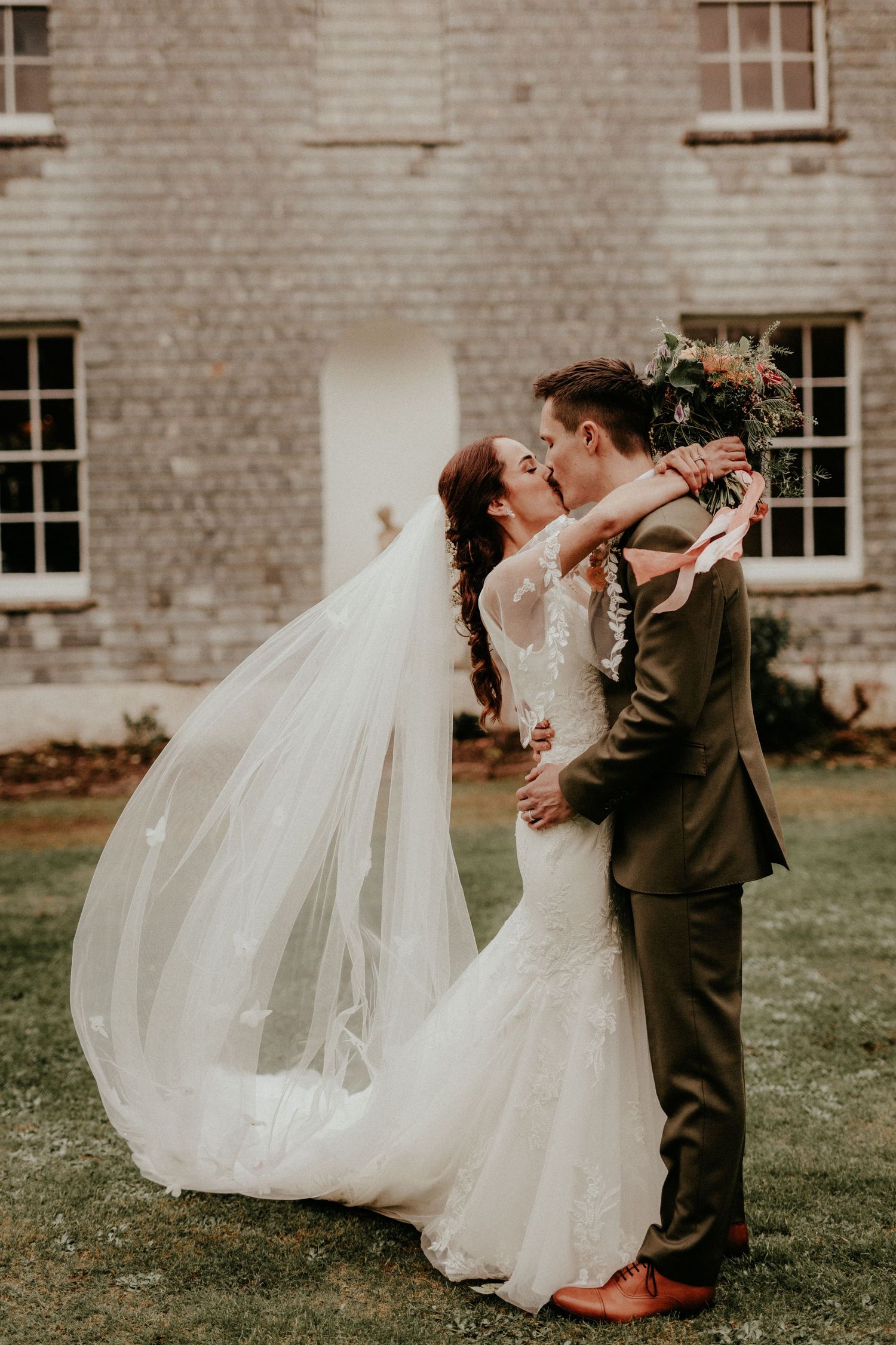 A romantic wedding couple embraces outdoors in front of a stone building as the bride's long veil flows in the breeze.