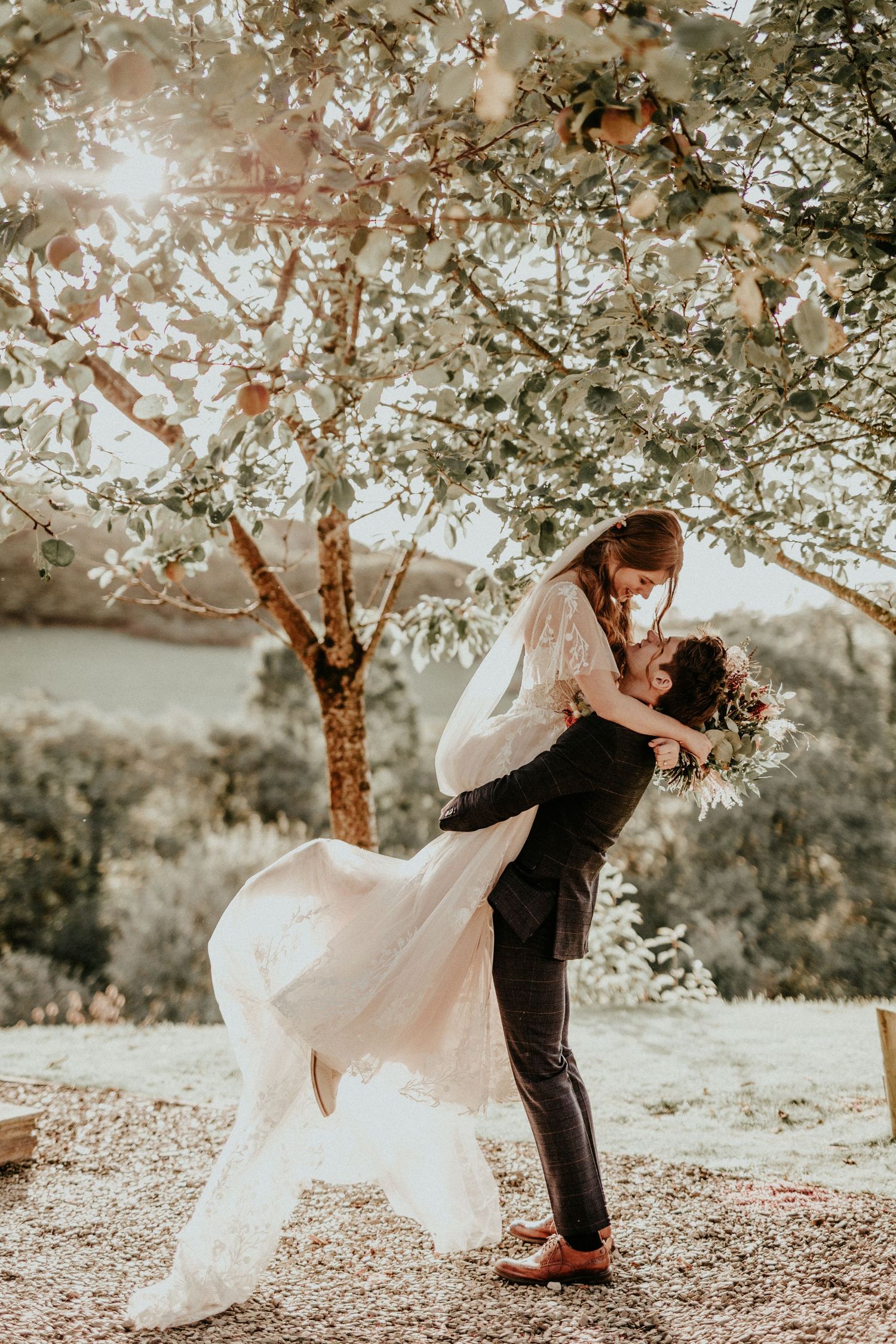 A romantic wedding couple embraces under a sunlit tree in an outdoor setting with flowing white dress and natural scenery.