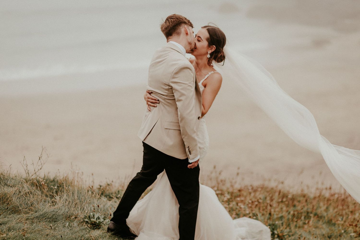 A loving couple embraces during their wedding photoshoot as the bride's veil flows dramatically in the mountain breeze.