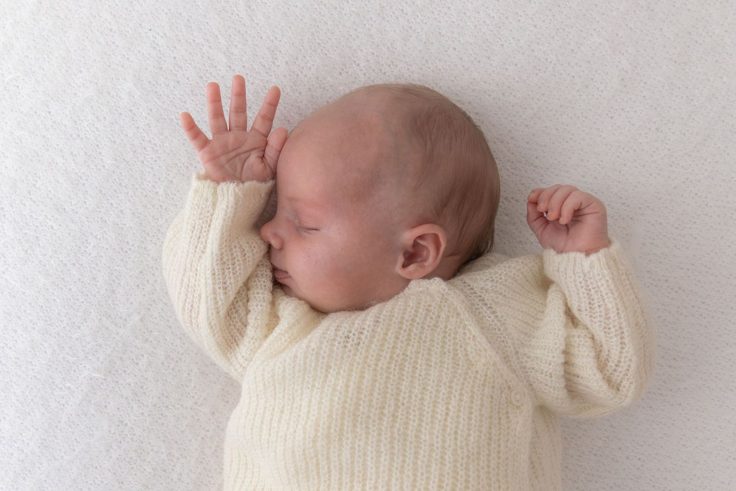 Sleeping baby wearing cozy cream knit sweater lying on white textured blanket.