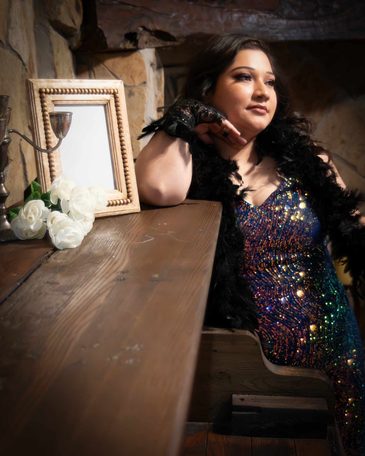 A person in a sparkly sequined dress poses elegantly near a vintage wooden table with a decorative frame and flowers.