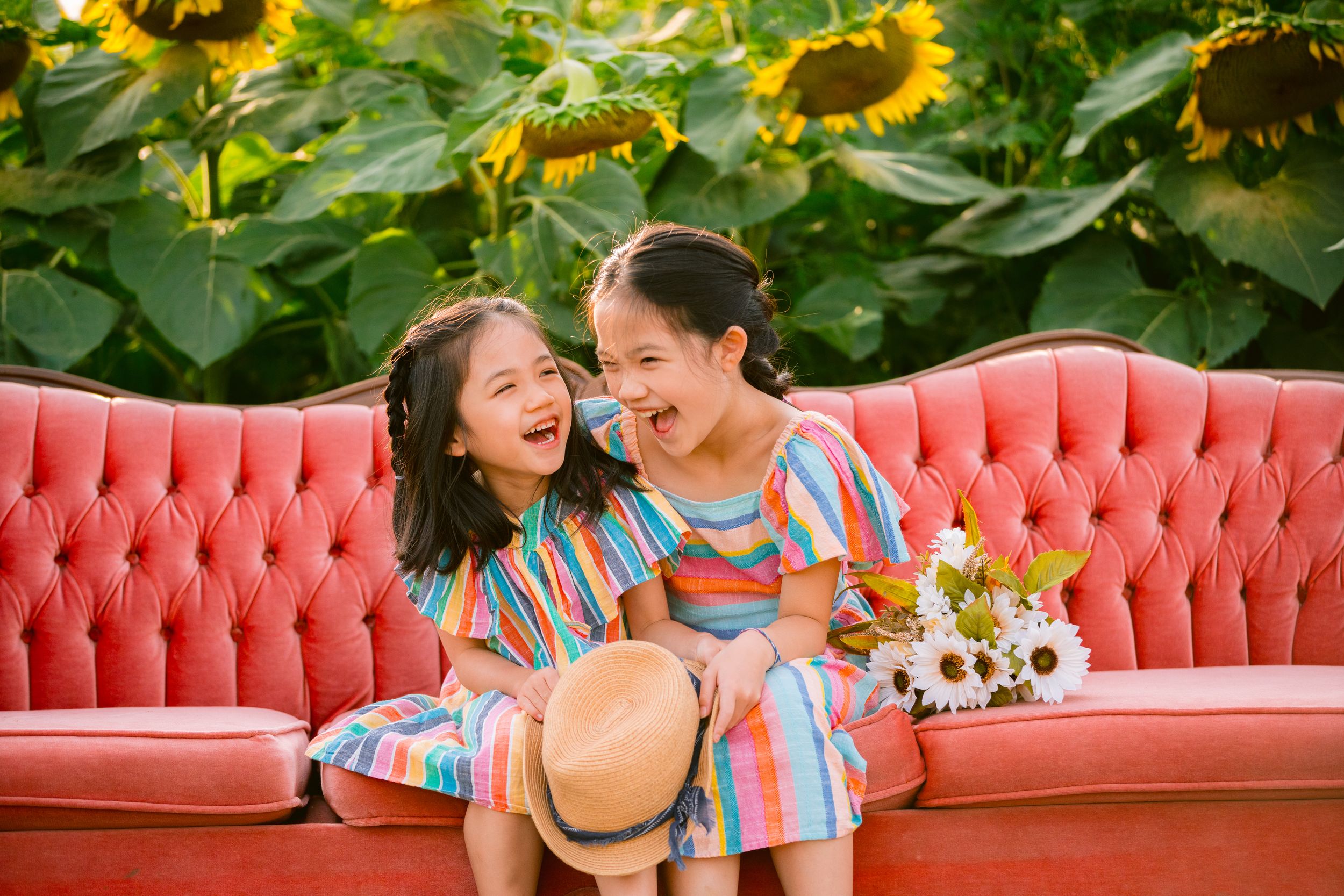 Two people share joyful laughter while sitting on a pink vintage couch with vibrant sunflowers blooming in the background.