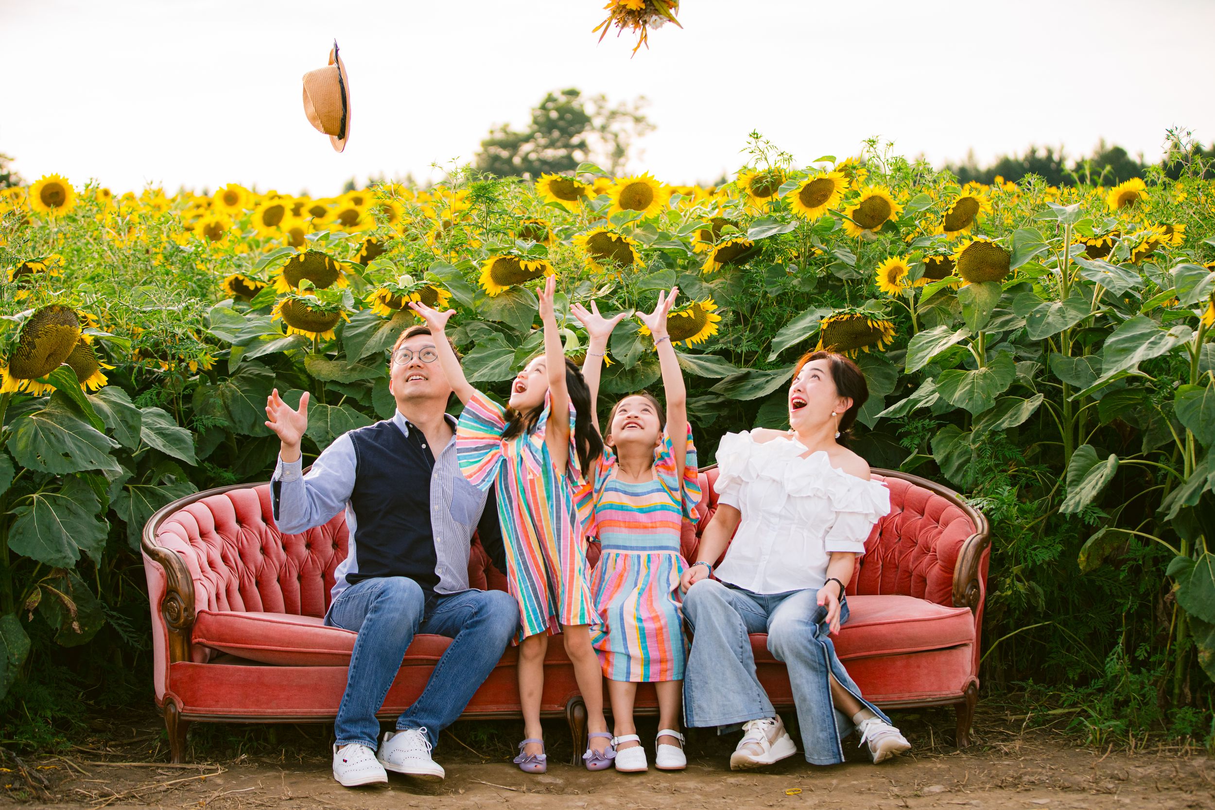 A vintage red couch sits in a vibrant sunflower field as playful figures toss leaves in late afternoon sunlight.