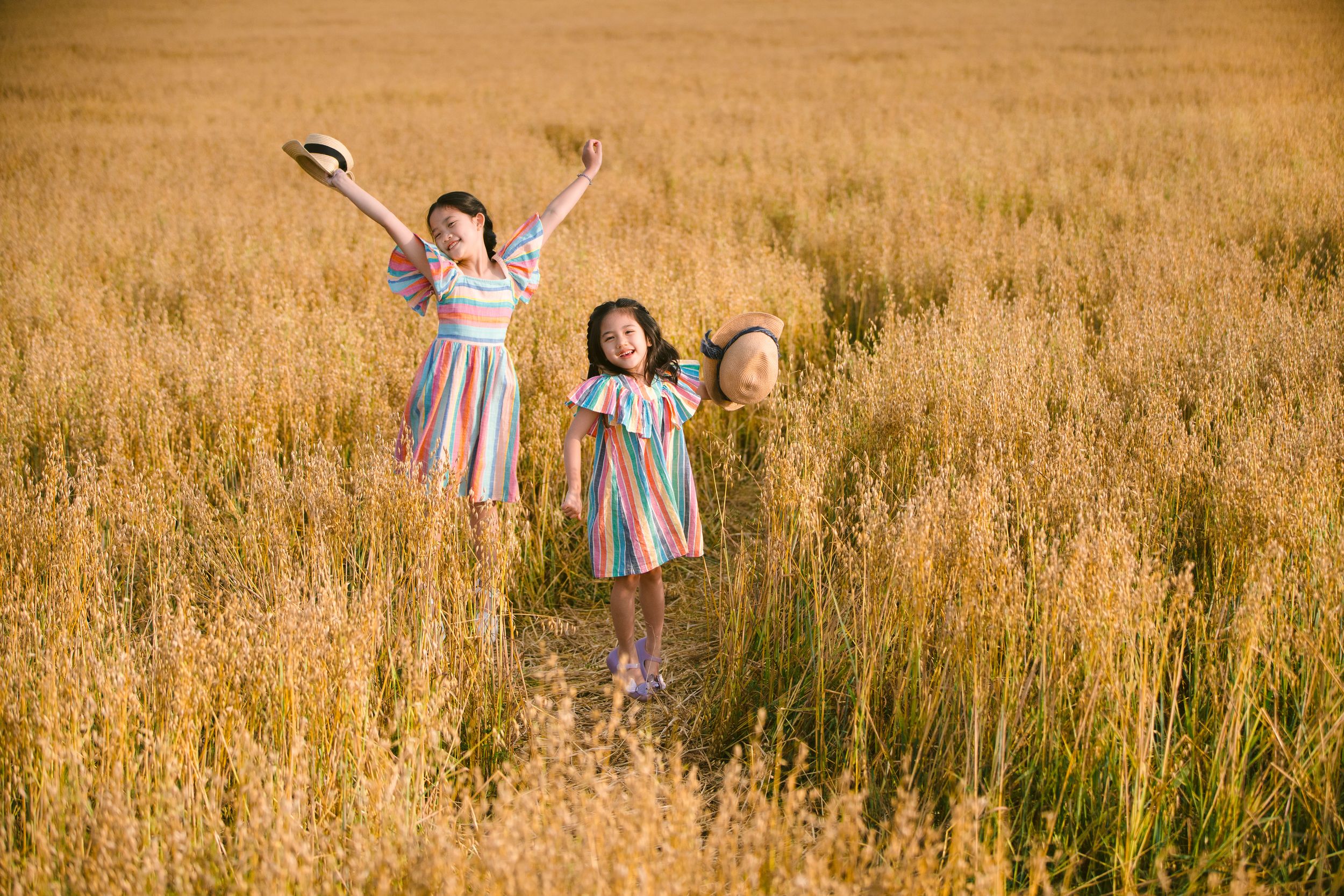 Two young children in dresses playing joyfully in a golden wheat field at sunset.