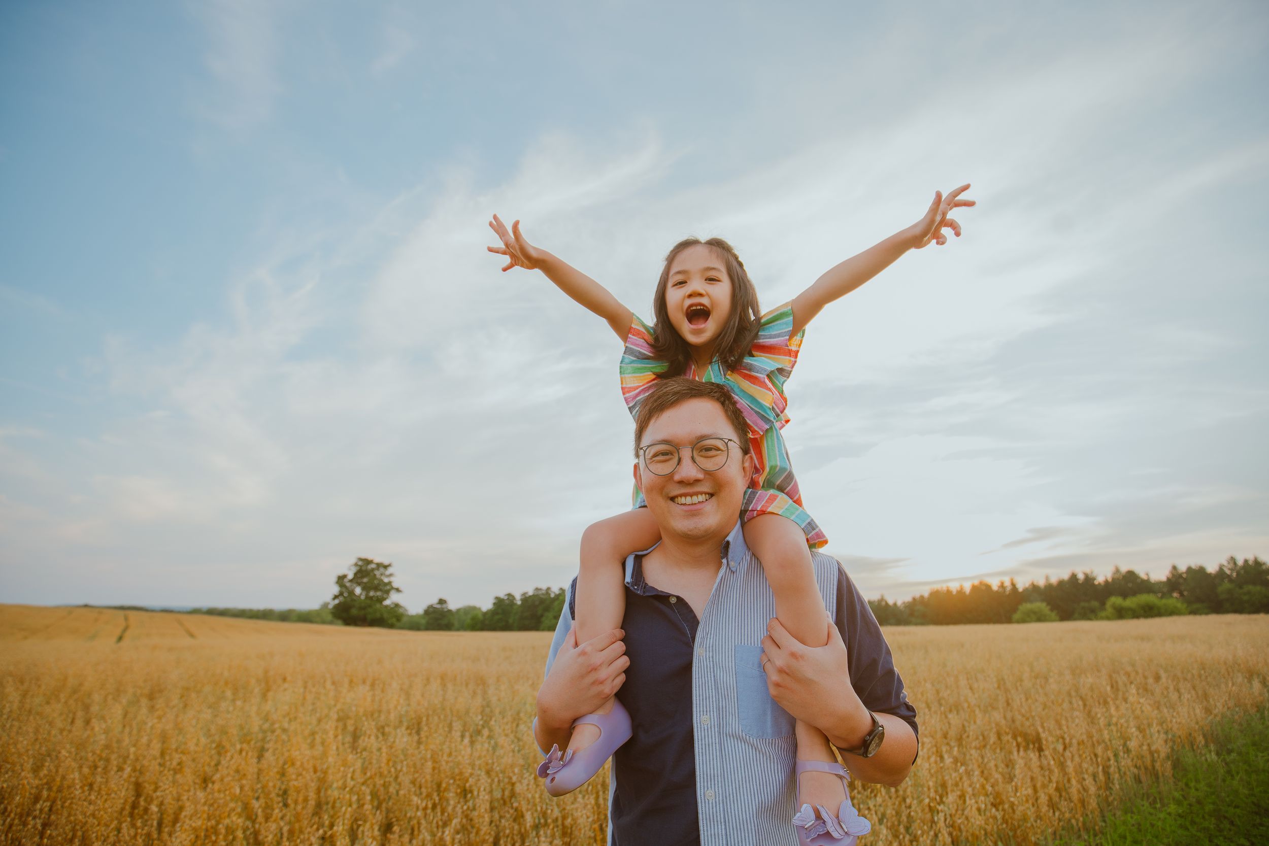 Two people joyfully playing in a golden wheat field during sunset with arms raised against a blue sky.