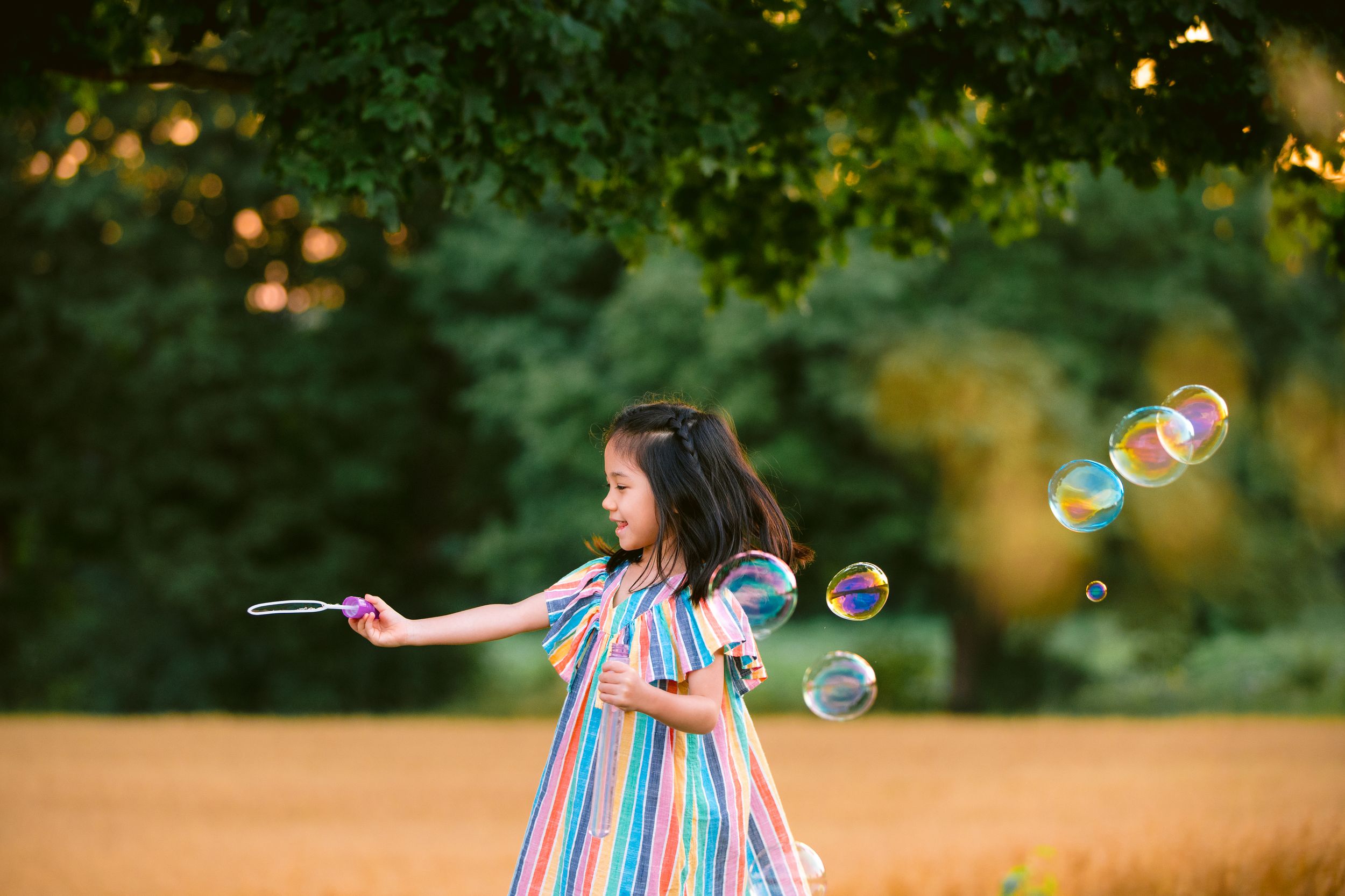 A young girl in a striped dress plays with bubbles outdoors during sunset.