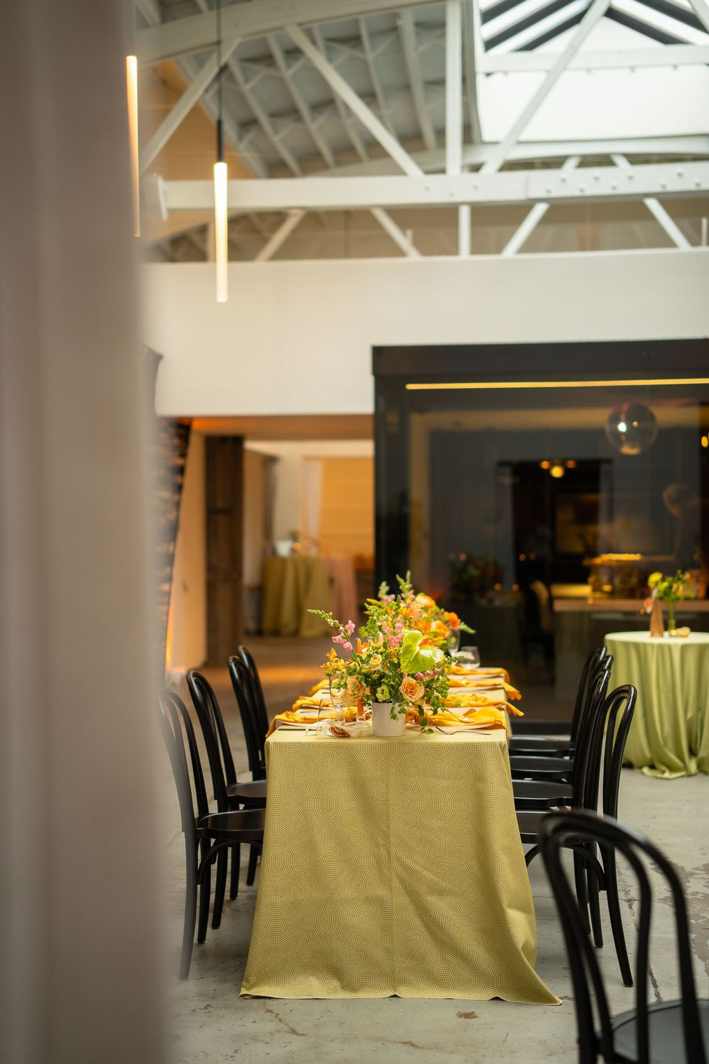 Long dining table with gold tablecloth and floral centerpieces set in an industrial-style event space.