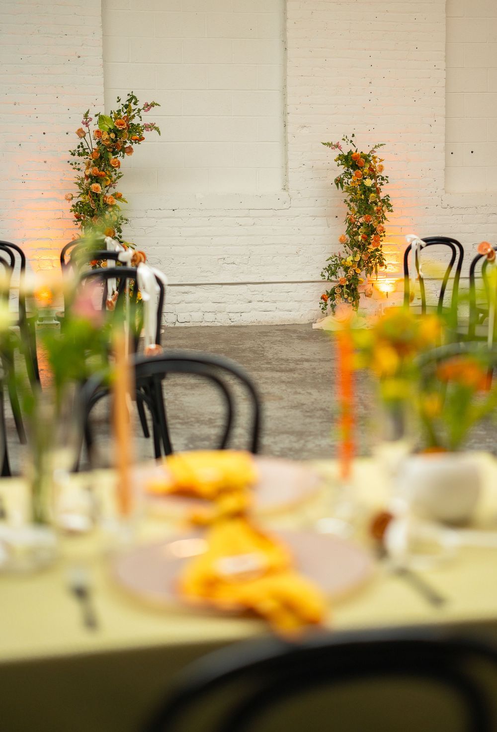 Table setting with yellow accents and floral arrangements against white brick wall.