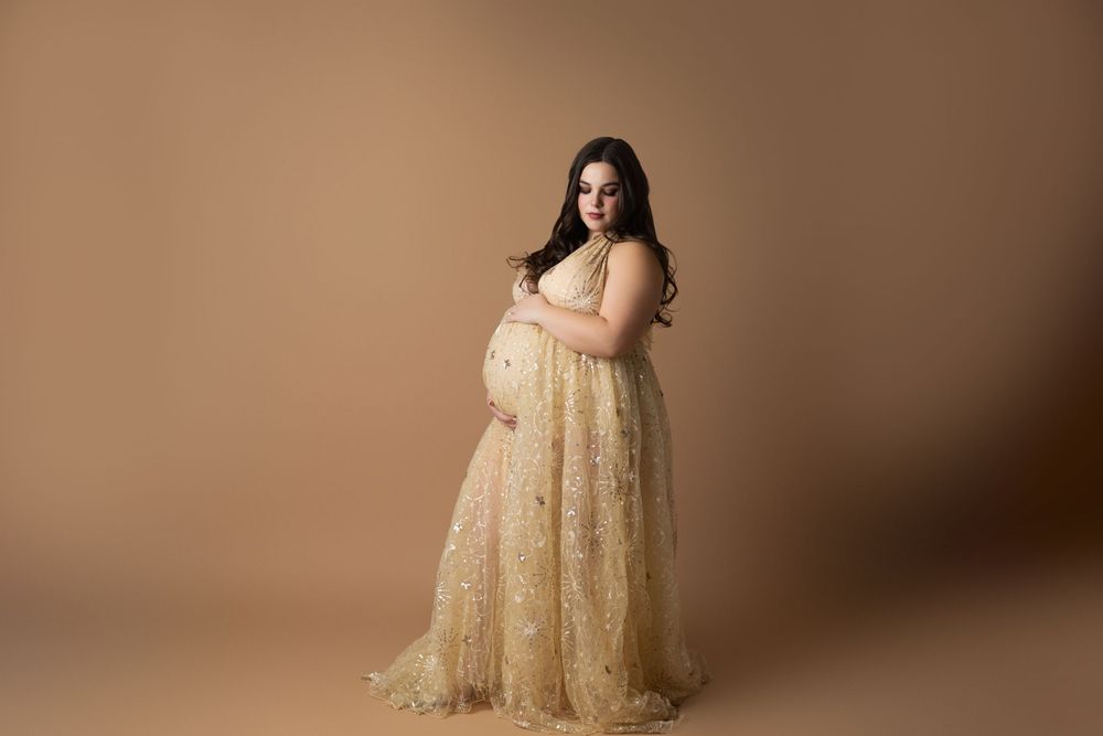 Expectant mother in a flowing champagne lace gown poses against a neutral brown backdrop during maternity photoshoot.