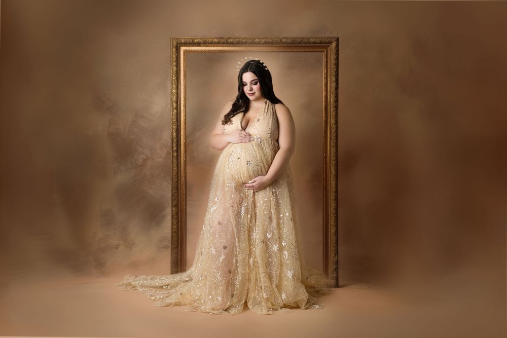 A pregnant woman poses in an elegant cream lace gown against a sepia backdrop with a vintage wooden frame.