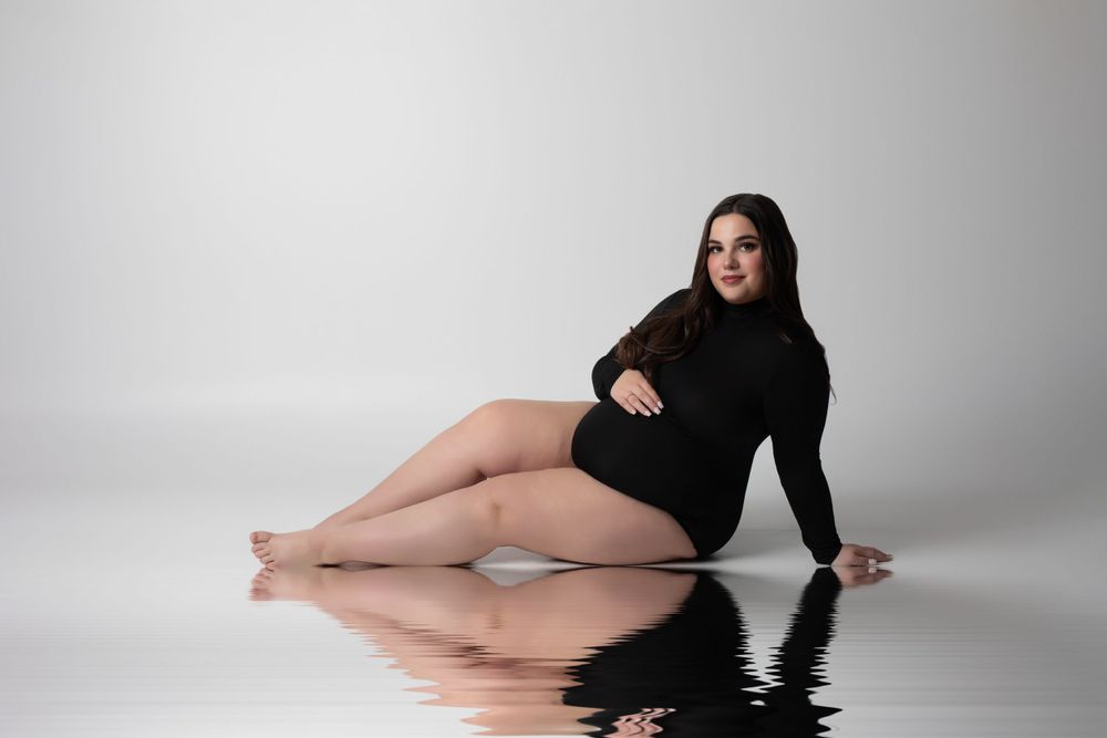 Model wearing black leotard poses elegantly on reflective floor in minimalist studio setting.