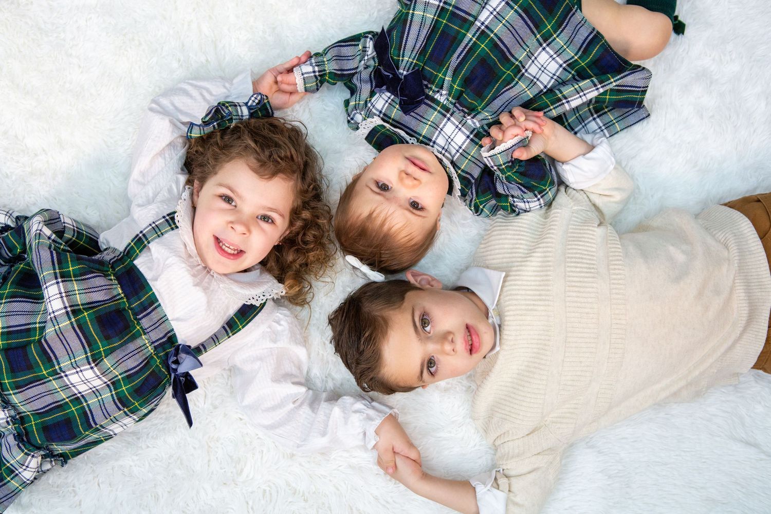 Three children lying in snow wearing plaid clothing and smiling at camera.