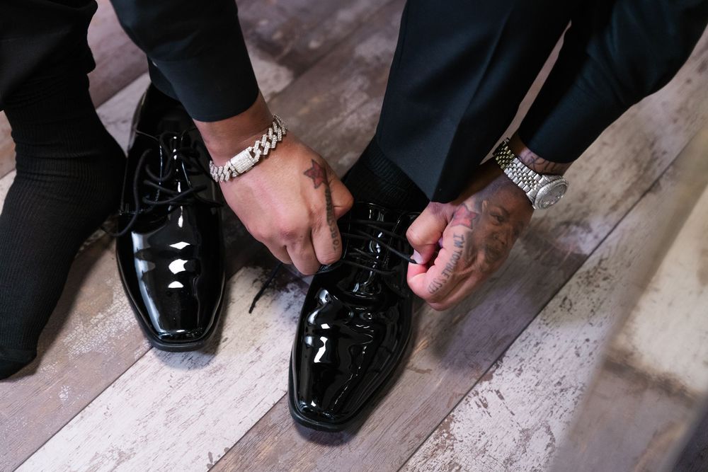 Person tying the laces on shiny black formal dress shoes while sitting on wooden floor.
